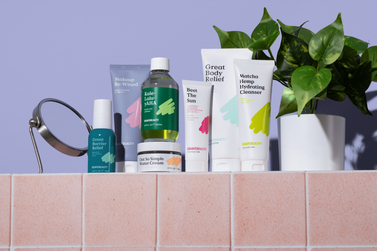 Krave beauty products on top of a peach tile table on the right side of the products there's a plant with a white pot on the left there's mirror and a purple background