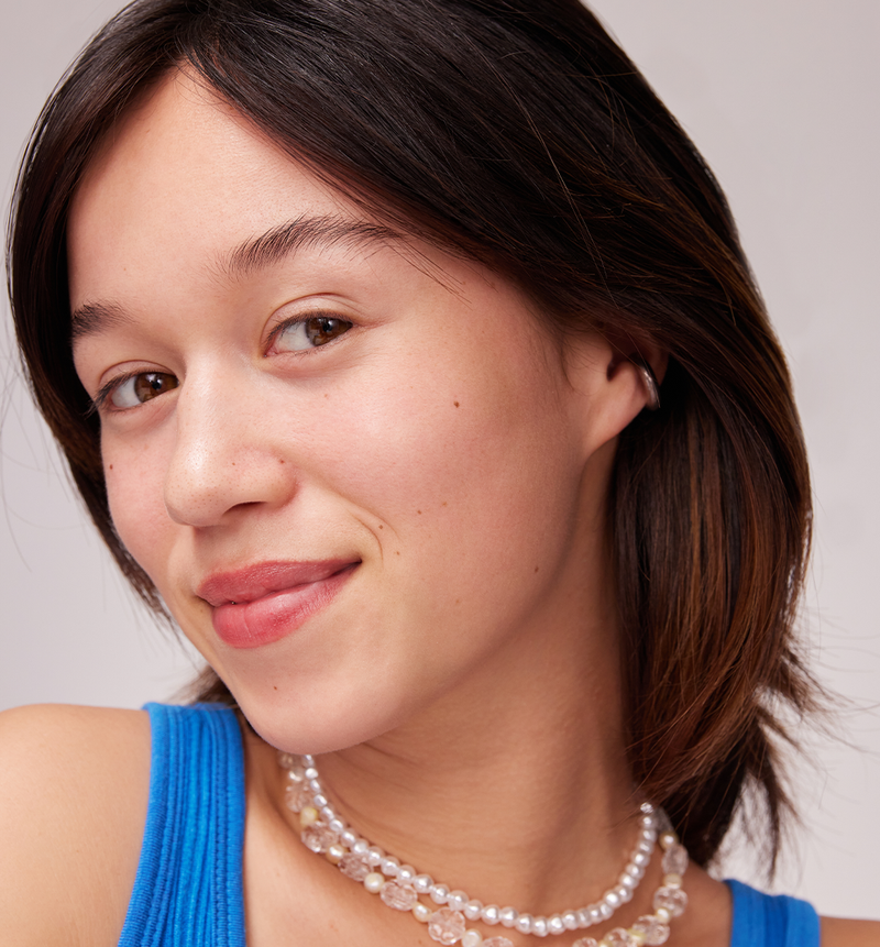 Woman with brown hair with a small smile, one white beed collar and another white / transparent collar and a blue tank top